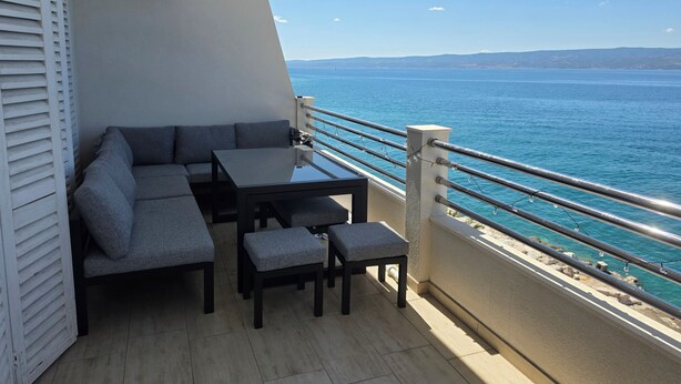 A balcony with a sofa, table, and chairs, overlooking a body of water. - ​First-Line Sea View: Luxurious 3-Room Family Apartment – 10 meters from the Water (Jesenice)