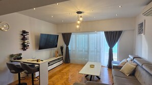 A modern living room with a sofa, a coffee table, a TV mounted on the wall, and a dining area with a table and chairs. - ​First-Line Sea View: Luxurious 3-Room Family Apartment – 10 meters from the Water (Jesenice)