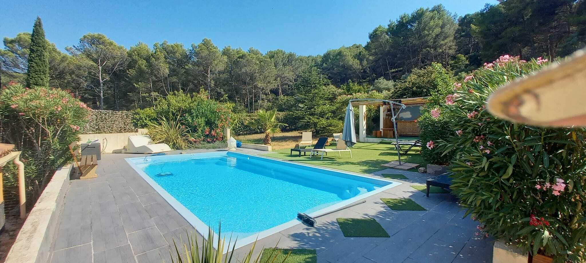 Una piscina rectangular con solárium empedrado, rodeada de follaje y un pequeño edificio con techo.
