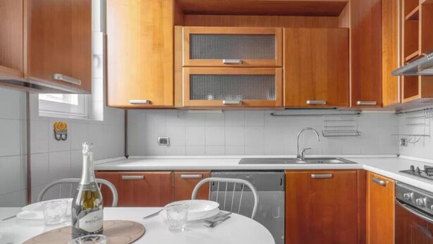 A modern kitchen with wooden cabinets, a white countertop, and a dining area set for two. - RI2 [Loreto-Cathedral] • Central • Large • (Milano)