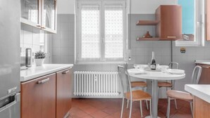 A kitchen with a white countertop, a round dining table with chairs, a window with curtains, and a radiator. - RI2 [Loreto-Cathedral] • Central • Large • (Milano)