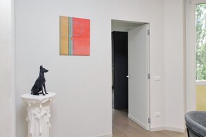 A hallway with a white wall, a black dog sculpture on a pedestal, a door leading to another room, and a window with a view of trees. - 4BNB - Luxury Chinotto Apartment (Roma)
