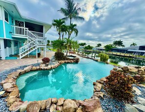 A swimming pool with a stone border, palm trees, and a building with a white balcony. - Tropical canal home - private pool, Fort Myers Beach, dock, direct gulf access (Fort Myers Beach)