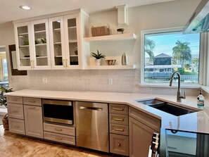 A modern kitchen with a stainless steel dishwasher, white cabinets, a window with a view of palm trees, and a built-in oven. - Tropical canal home - private pool, Fort Myers Beach, dock, direct gulf access (Fort Myers Beach)