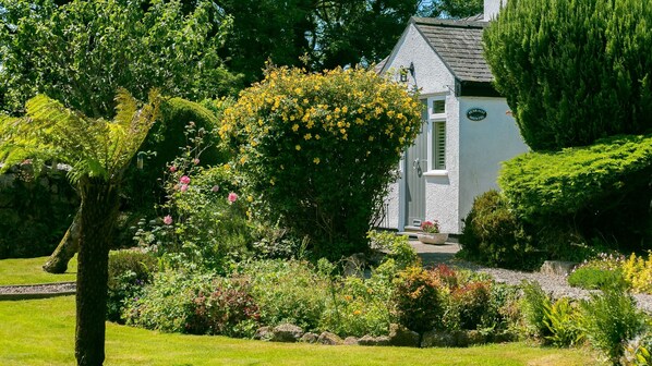 Un bâtiment blanc surmonté d’un toit gris, entouré d’une végétation luxuriante et de plantes en fleurs.