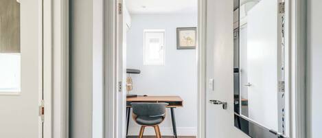 A hallway leading to a room with a desk, chair, and framed picture on the wall.