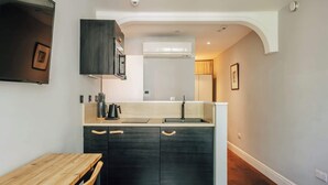 A compact kitchen with dark cabinetry, a wooden dining table, and a built-in air conditioning unit. - The Knaresborough -Stylish Earl's Court Apartment (London)
