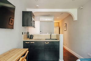 A compact kitchen with dark cabinetry, a wooden dining table, and a built-in air conditioning unit. - The Knaresborough -Stylish Earl's Court Apartment (London)