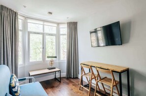 Ein Zimmer mit großem Fenster, einem flachbildfernseher an der Wand, einem Holztisch und einer Bank.