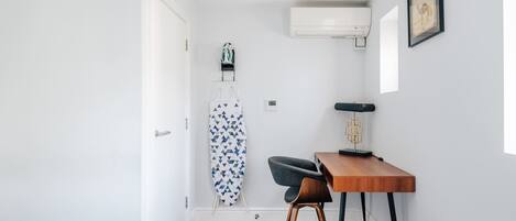 Un dormitorio con cama, escritorio, silla, tabla de planchar y aire acondicionado instalado en la pared.