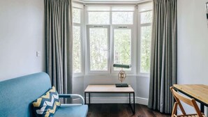 A room with a bay window, a blue sofa, a wooden table, and a lamp. - The Knaresborough -Stylish Earl's Court Apartment (London)