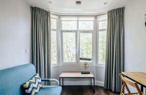 A room with a bay window, a blue sofa, a wooden table, and a lamp. - The Knaresborough -Stylish Earl's Court Apartment (London)