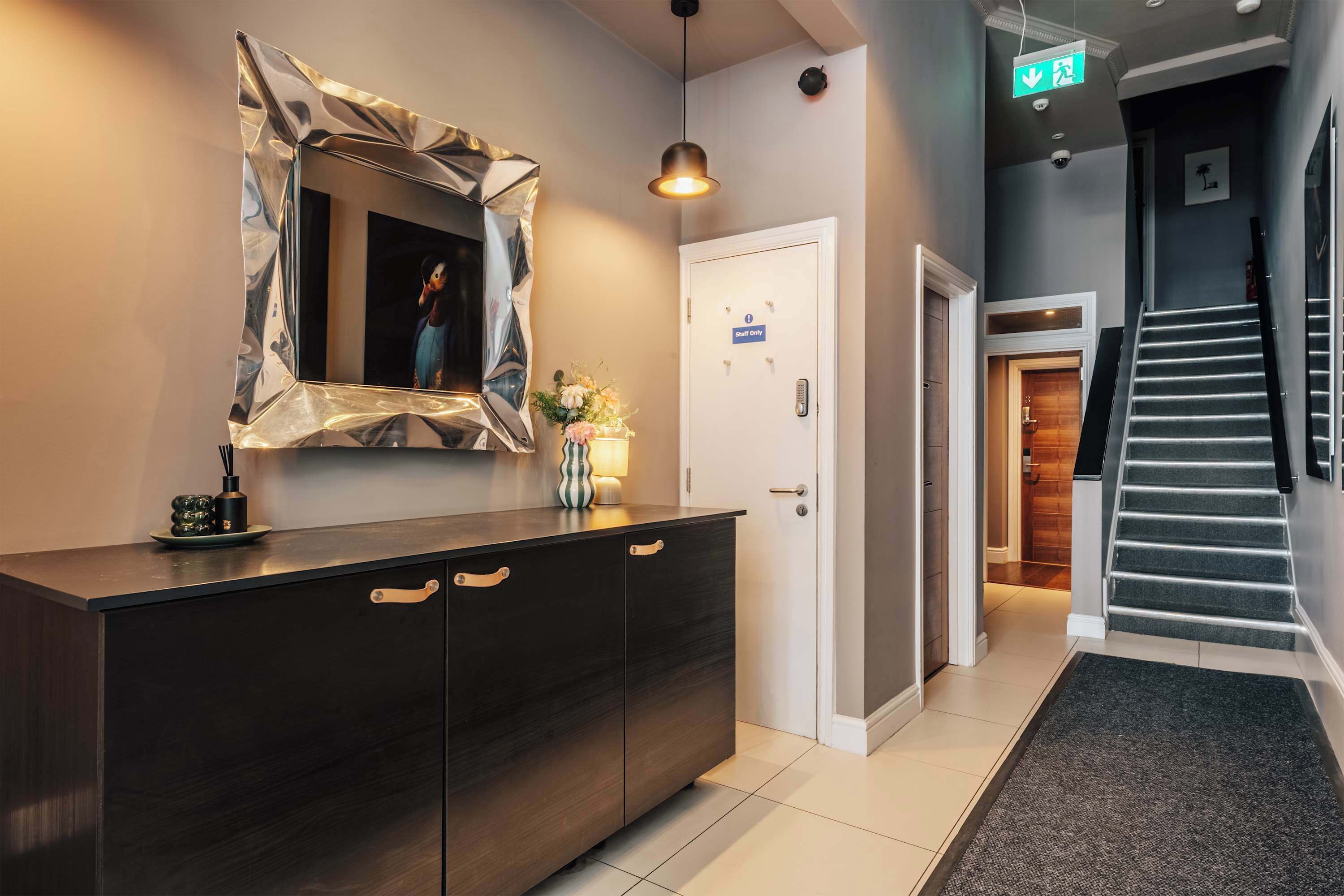 Un moderno vestíbulo de hotel con un mostrador de recepción de madera oscura, un espejo con marco geométrico, una escalera y una puerta con el letrero 'Staff Only'.