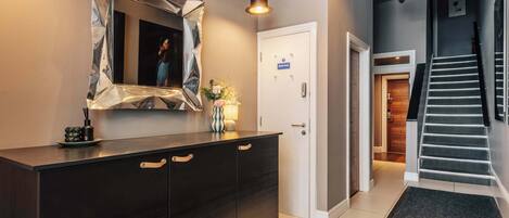 Un moderno vestíbulo de hotel con un mostrador de recepción de madera oscura, un espejo con marco geométrico, una escalera y una puerta con el letrero 'Staff Only'.