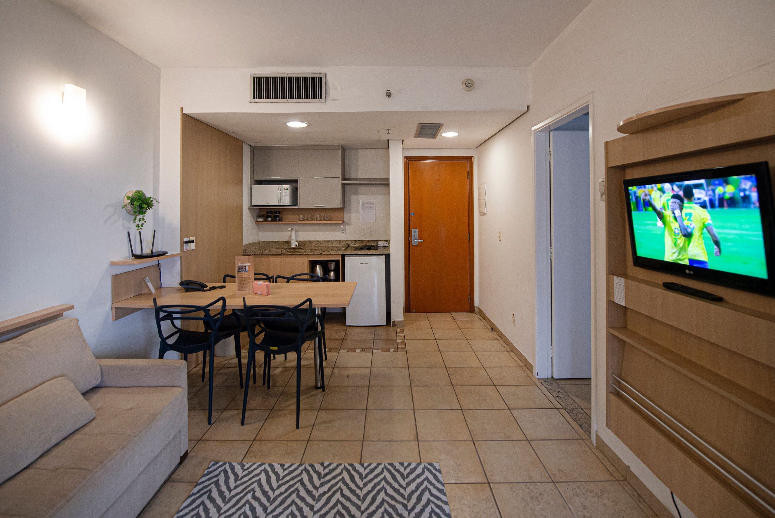 A modern living room with a sofa, dining table, chairs, a TV, and a kitchen in the background.