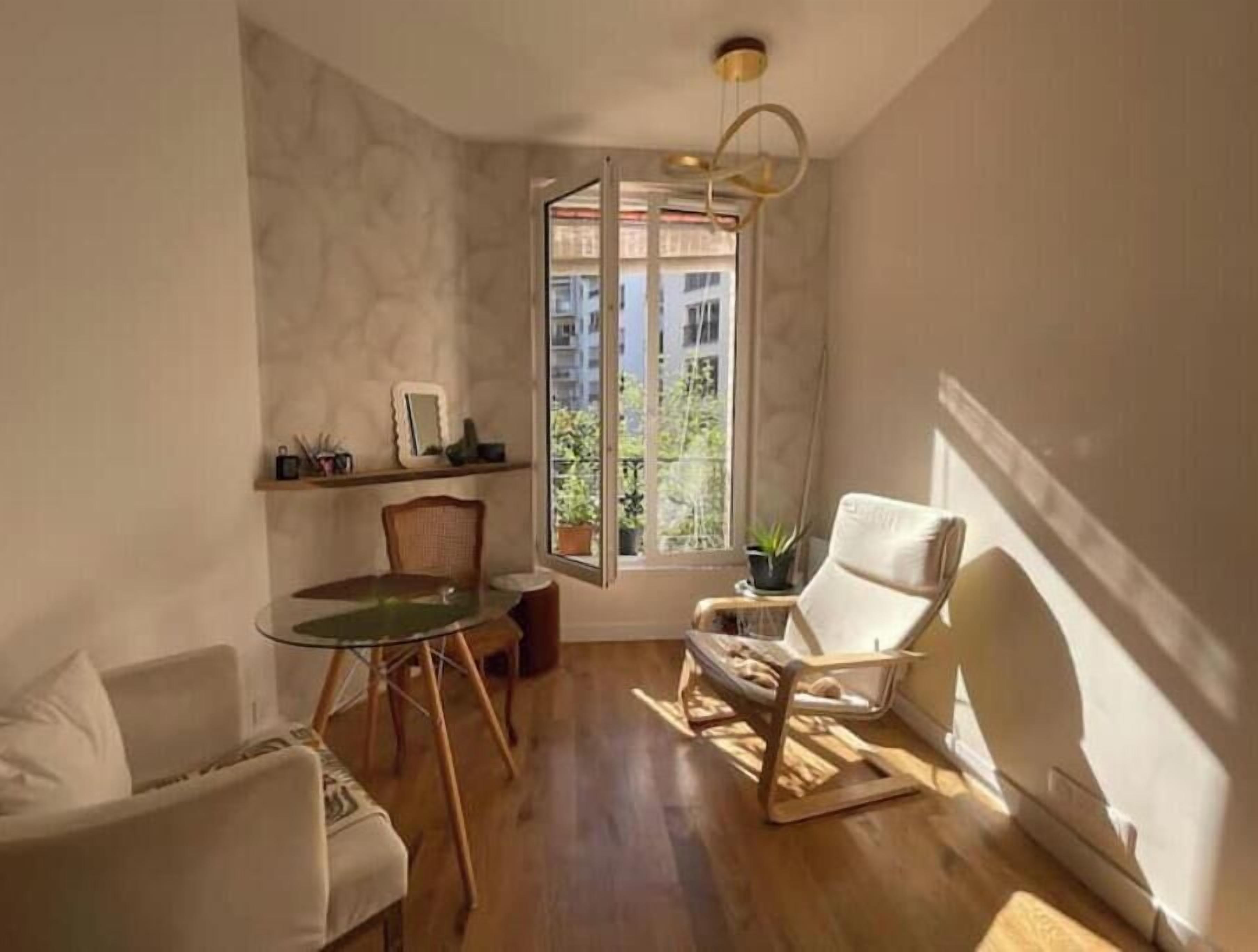 Een woonkamer met een glazen tafel, twee stoelen, een bank, een witte fauteuil, een klein bijzettafeltje met een plant en uitzicht op gebouwen door een groot raam.
