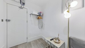 Ein Hotelzimmer mit einer weißen Tür, einem kleinen Tisch mit einer Fernbedienung, einem Handtuchhalter mit Handtüchern und einer Schachtel sowie einer wandmontierten Lampe.