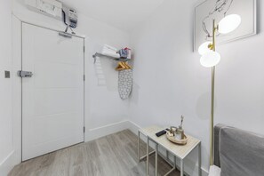 Ein Hotelzimmer mit einer weißen Tür, einem kleinen Tisch mit einer Fernbedienung, einem Handtuchhalter mit Handtüchern und einer Schachtel sowie einer wandmontierten Lampe.