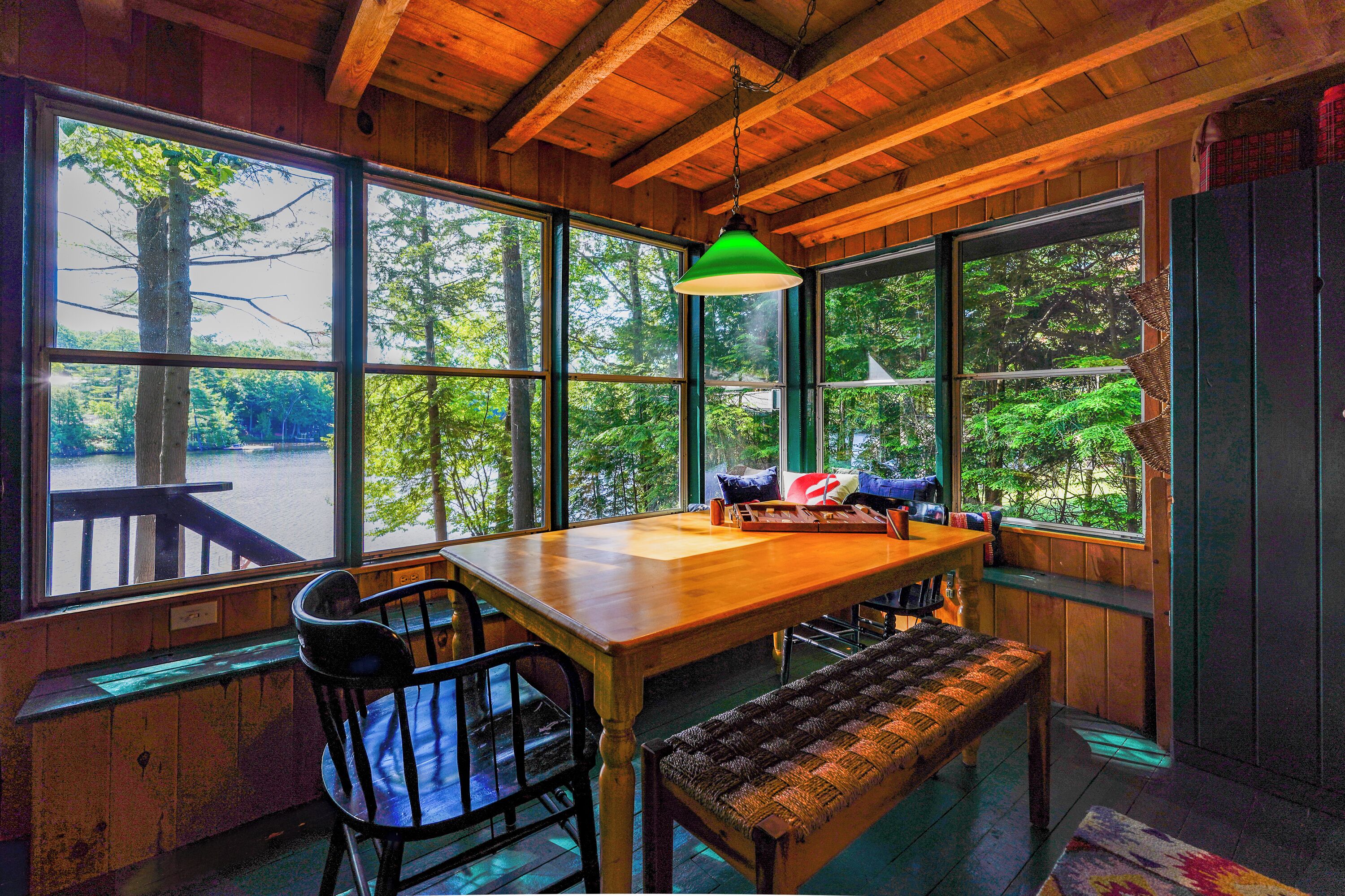 A wooden interior with a table, chairs, a bench, and large windows offering a view of trees and a body of water.