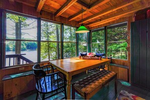 Un intérieur en bois comprenant une table, des chaises, un banc et de grandes fenêtres donnant sur des arbres et une étendue d’eau.