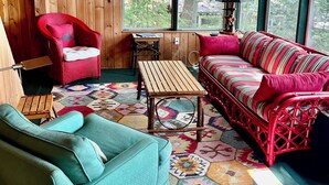 A cozy cabin interior with a wooden ceiling, a red wicker chair, a teal sofa, a wooden coffee table, and a colorful rug.