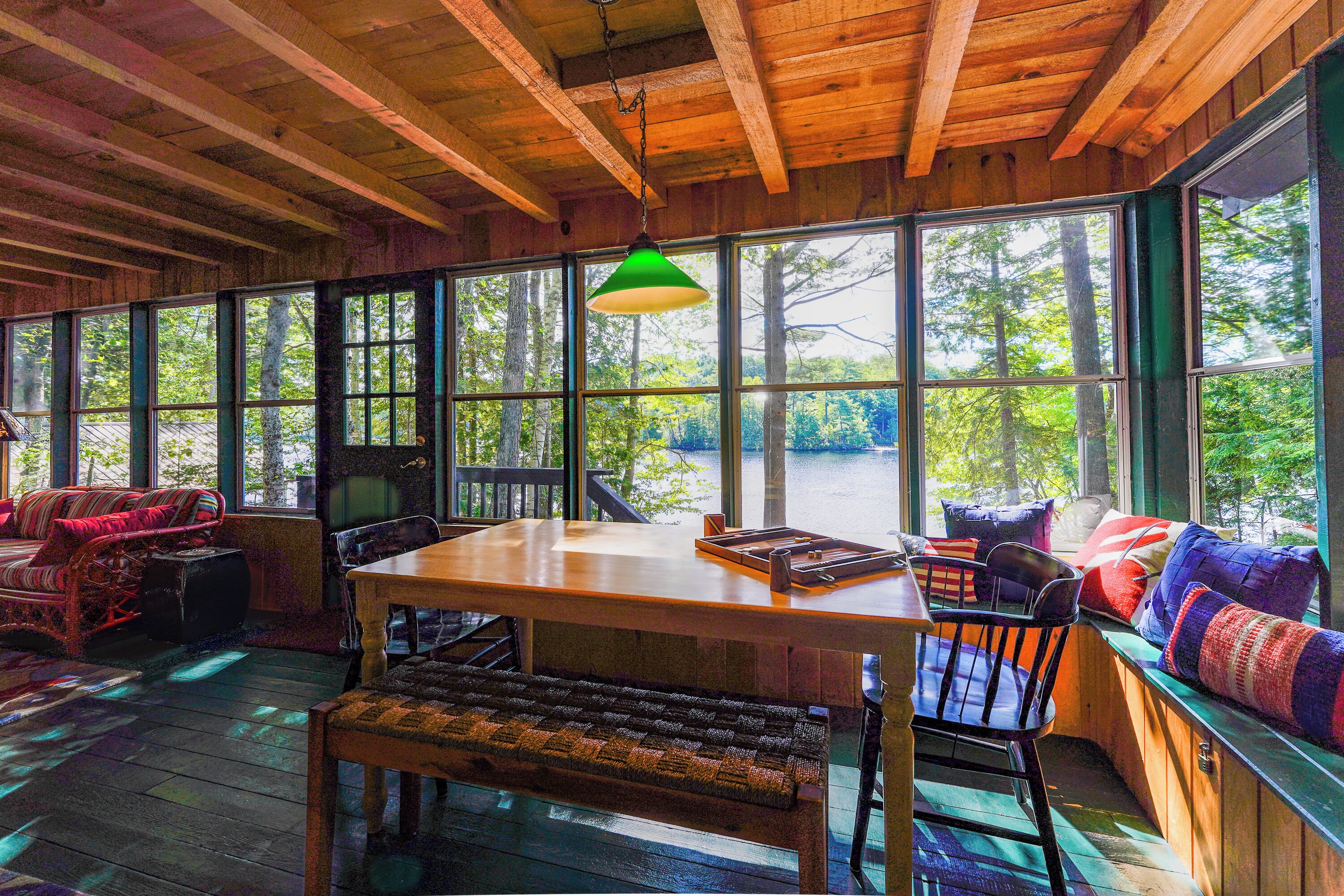 Interior de madera con una mesa de comedor, bancos y un sofá con cojines de colores. Amplias ventanas ofrecen vistas al exterior.