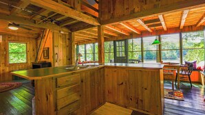 A wooden cabin interior with a large kitchen island, wooden cabinets, and large windows offering views of the outdoors.