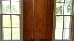 A room with a wooden paneled wall, a table with a white tablecloth, and two windows with a view of a grassy area.