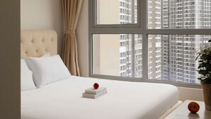 Habitación de hotel con una cama grande, una ventana con vistas a edificios altos, un cuadro en la pared y una planta en maceta sobre la mesita de noche.