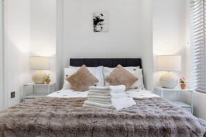 A neatly made bed with a fluffy blanket, two bedside tables with lamps, and a wall-mounted artwork. - Harlesden Comfort Studio (London)