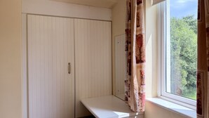 Ein Zimmer mit weißem Schreibtisch, einer Sitzbank, einem Kleiderschrank und einem Fenster mit Vorhängen.