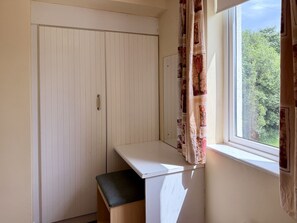 Ein Zimmer mit weißem Schreibtisch, einer Sitzbank, einem Kleiderschrank und einem Fenster mit Vorhängen.