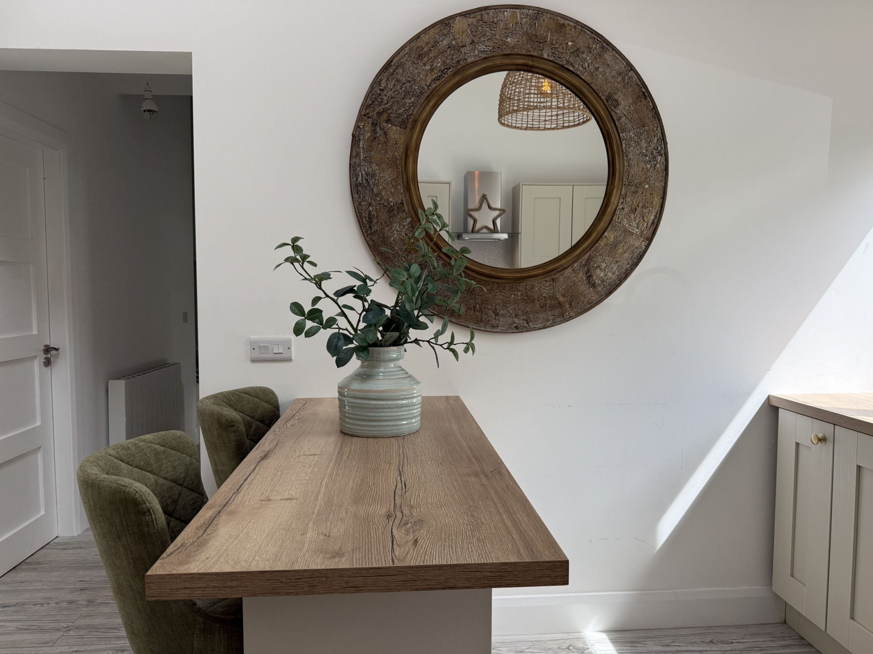 A room with a wooden table, a green chair, a vase with a plant, and a round mirror on the wall.