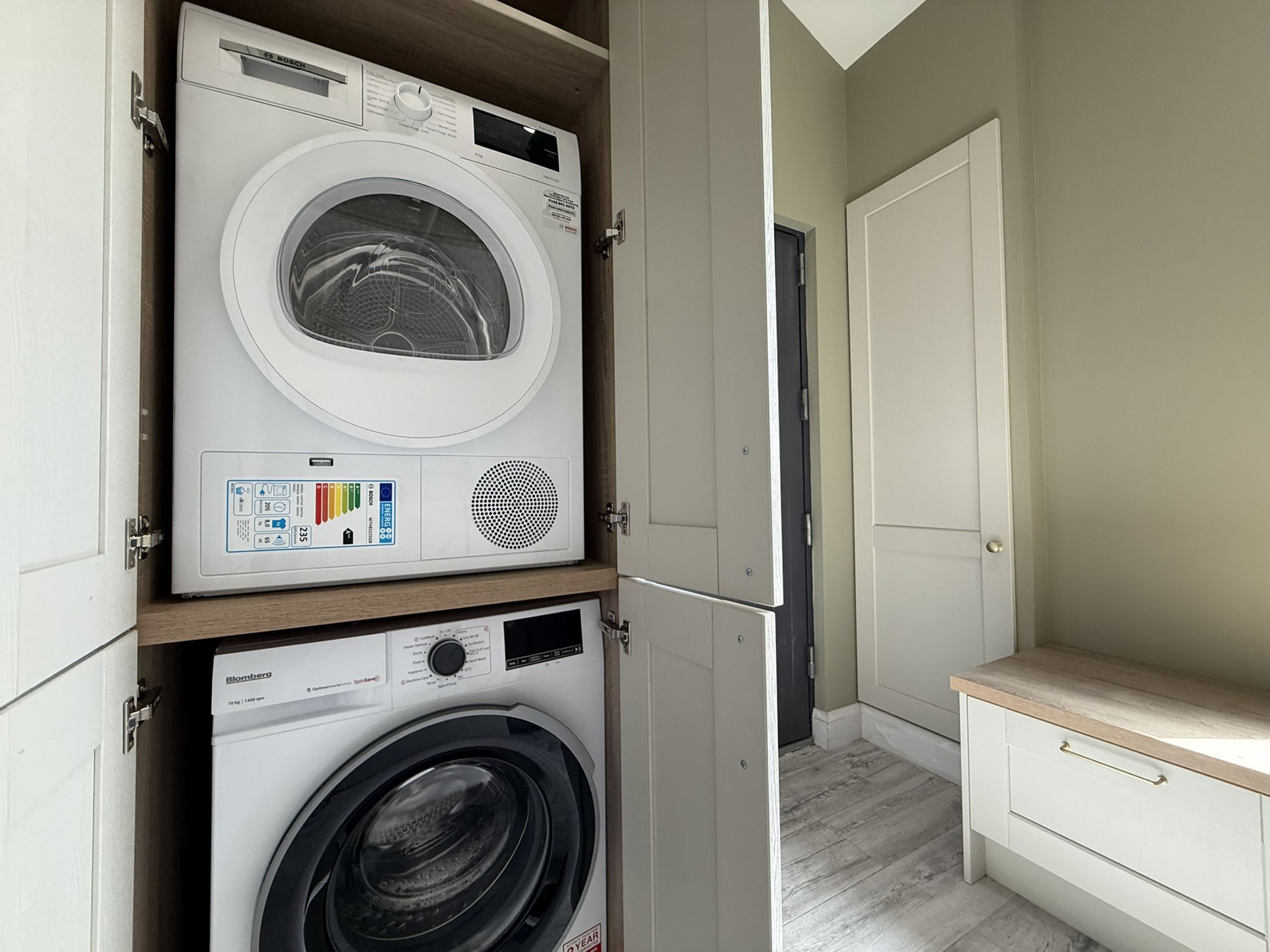A laundry room with two stacked washing machines, one Bosch and one Blomberg, and a wooden bench.
