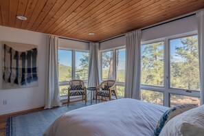 A bedroom with a large bed, two chairs, a small table, and a view of trees through large windows. - Mountain Retreat in the Sangre de Cristo Foothills (Santa Fe)