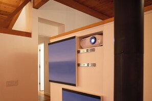 Een kamer met een houten plafond, een witte muur met een blauw paneel, een zwarte radiator en een projector aan het plafond.