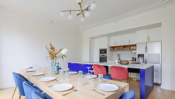 A modern kitchen with white cabinets, a central island, and a dining area with a wooden table and blue chairs. - Luxurious Apartment - 4br/10p- Ternes/pereire (Paris)