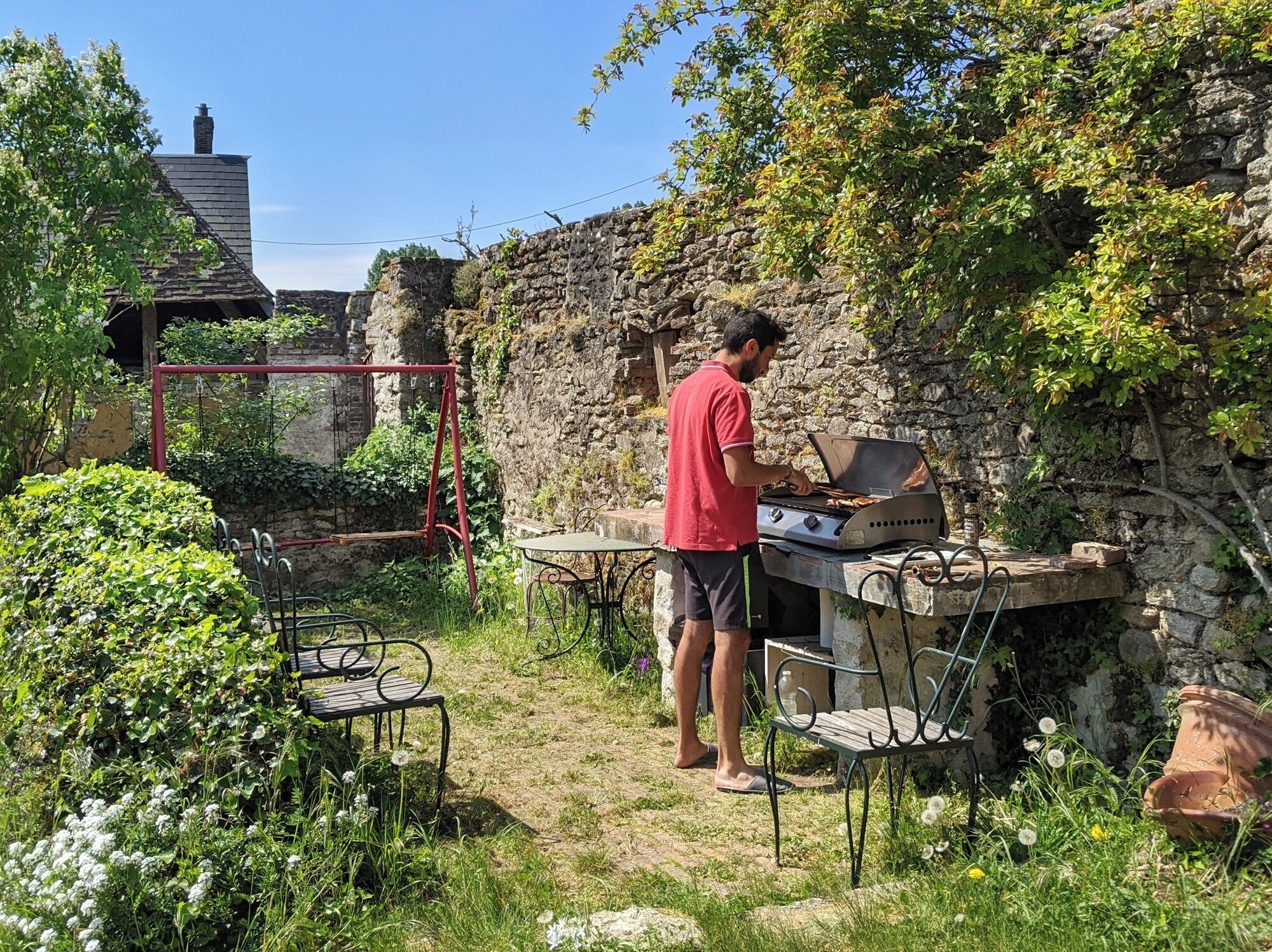 Un homme qui fait griller de la viande à l’extérieur, avec un mur de pierre et de la verdure en arrière-plan.