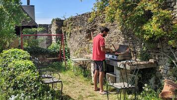Un homme qui fait griller de la viande à l’extérieur, avec un mur de pierre et de la verdure en arrière-plan.