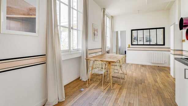 A room with a wooden floor, a dining table with chairs, large windows with curtains, and a radiator. - Modern Flat - 2ch/6p - Bastille (Paris)