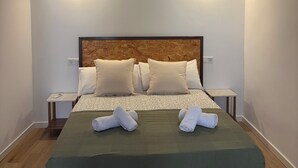 A neatly made bed with pillows, a green bedspread, and two rolled towels on the bed. There are two bedside tables with lamps, a wooden headboard, and a wooden ceiling beam.
