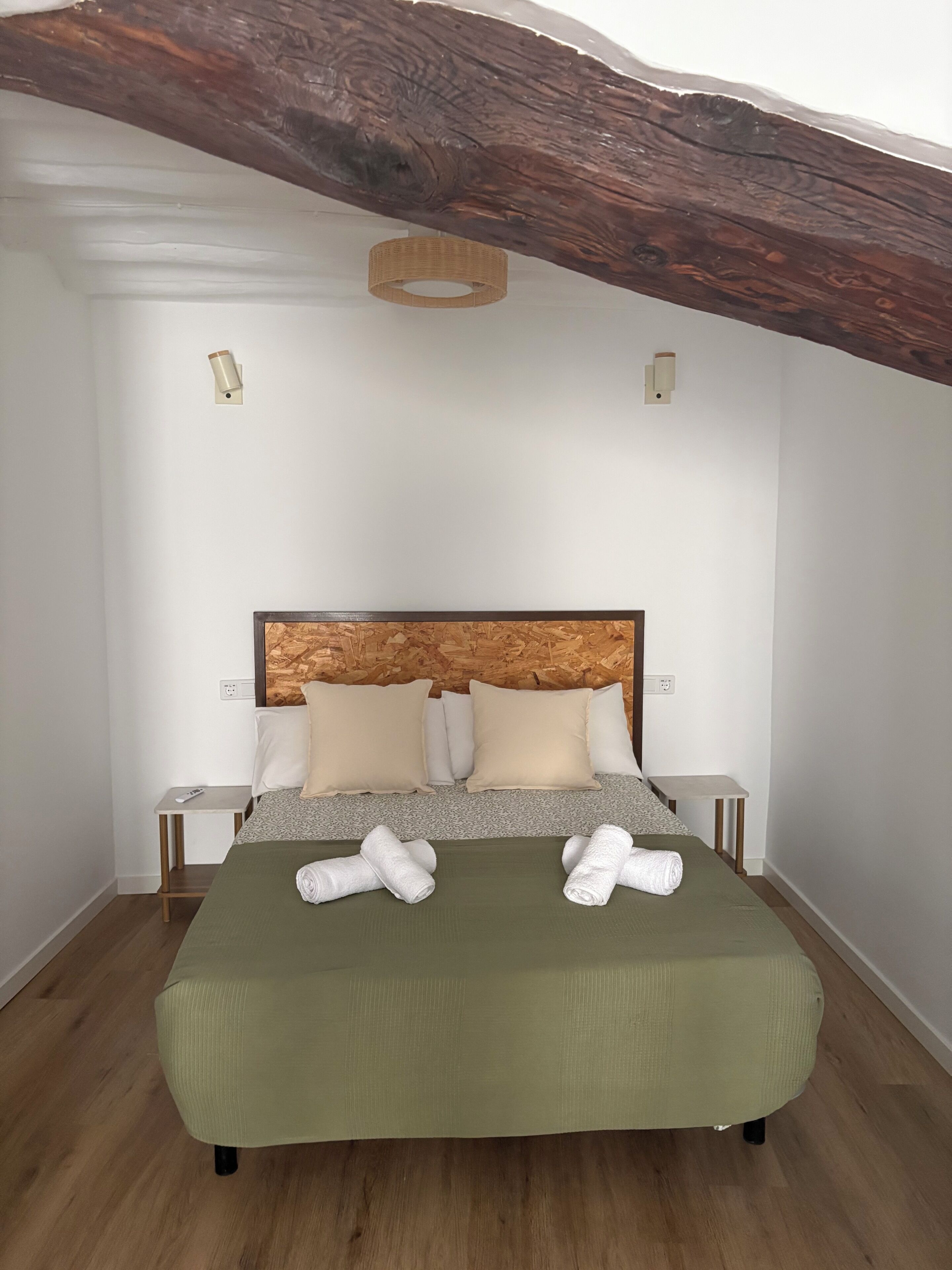 Een slaapkamer met een houten hoofdbord, twee nachtkastjes, een bed met een groene spreideken en twee opgerolde handdoeken op het bed.