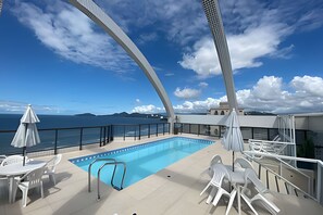 A pool area with a clear view of the sea and mountains, surrounded by white chairs and umbrellas. - SHB Melhor prédio com vista para o mar (Santos)