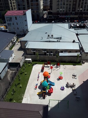 Vista aérea de un parque infantil con tobogán y estructuras para escalar, rodeado de edificios y un estacionamiento.