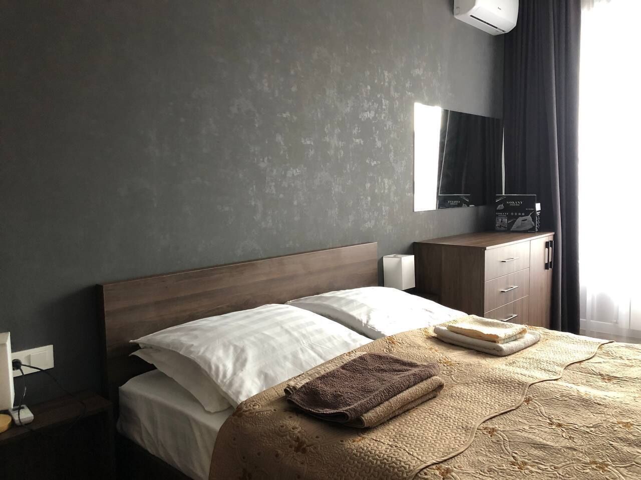 A hotel room with a bed, a wooden headboard, a nightstand, a lamp, a dresser, and a mirror.