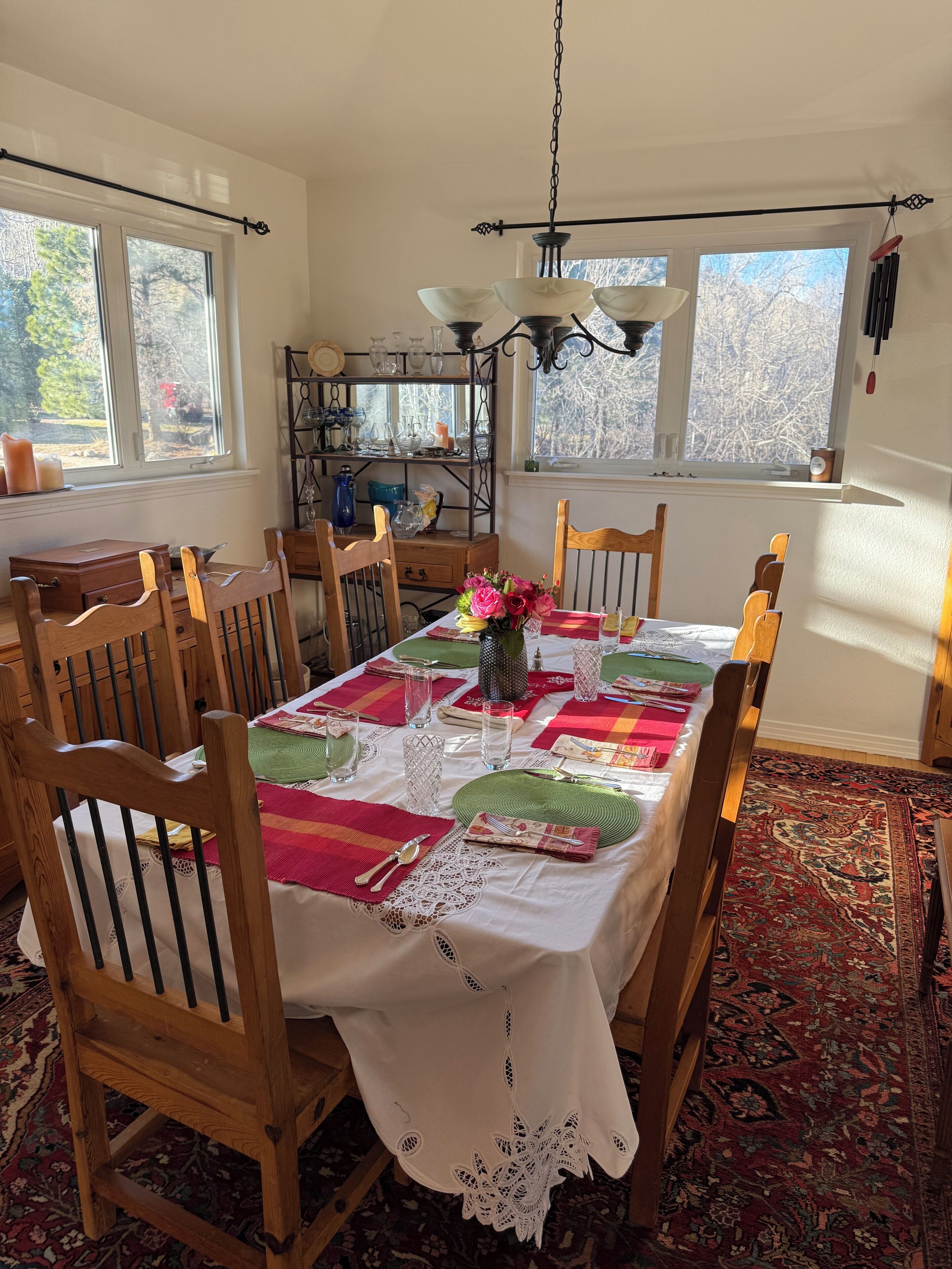 A dining room with a table set for six, surrounded by wooden chairs, a lace table runner, and a patterned rug.