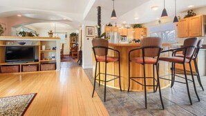 Una cocina moderna con armarios de madera, una isla central con taburetes altos y un comedor con televisor y estanterías.