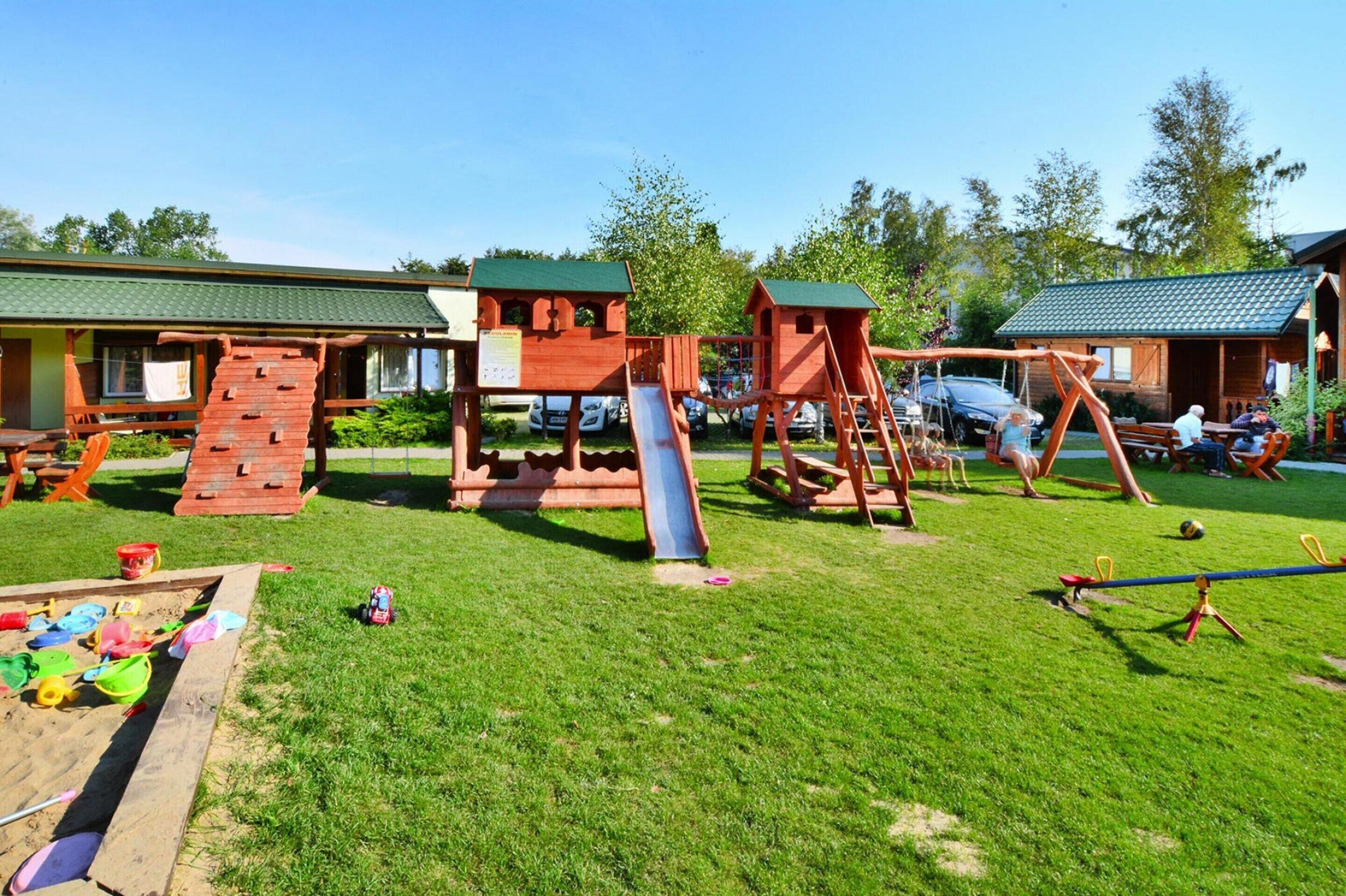 A playground with a slide and climbing structures, surrounded by a grassy area and wooden buildings.