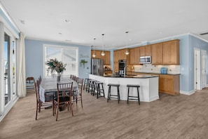 A modern kitchen with wooden cabinets, a central island with stools, and a dining area with a table and chairs. - 5BR Heated pool Oct-Nov, sunset views, near beach (Carolina Beach)
