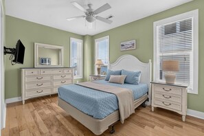 Un dormitorio con cama, mesitas de noche, cómoda, ventilador de techo y ventana con persianas.